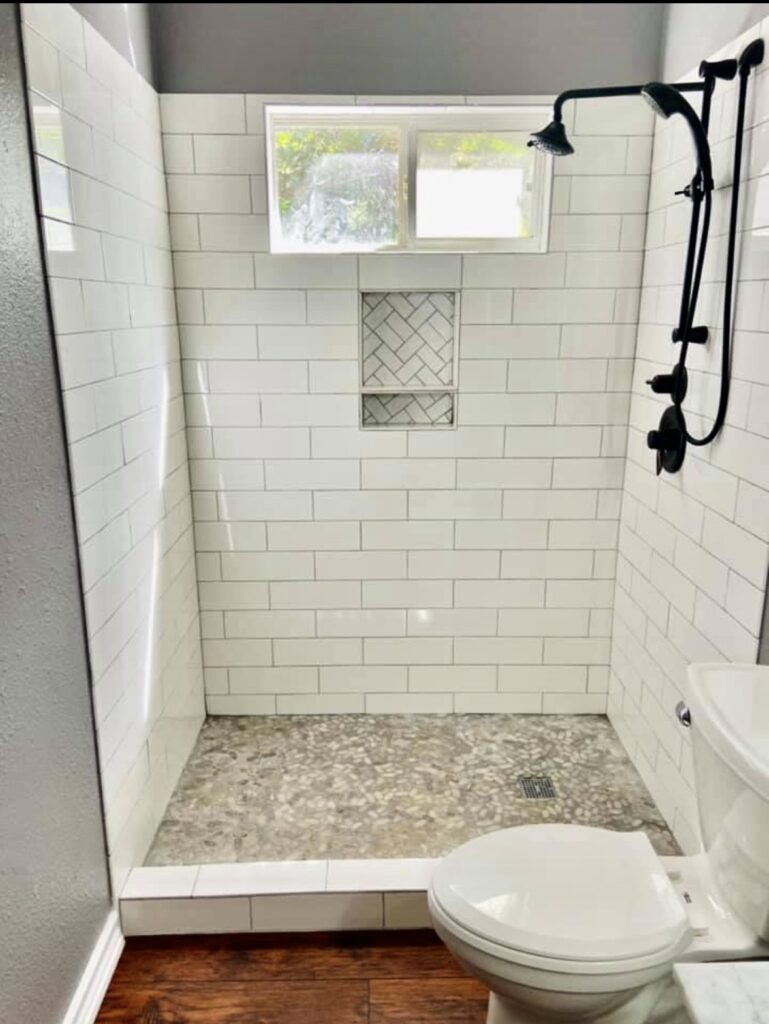 White tiled shower with a high window, recessed niche, dark fixtures and a mosaic floor beside a toilet.