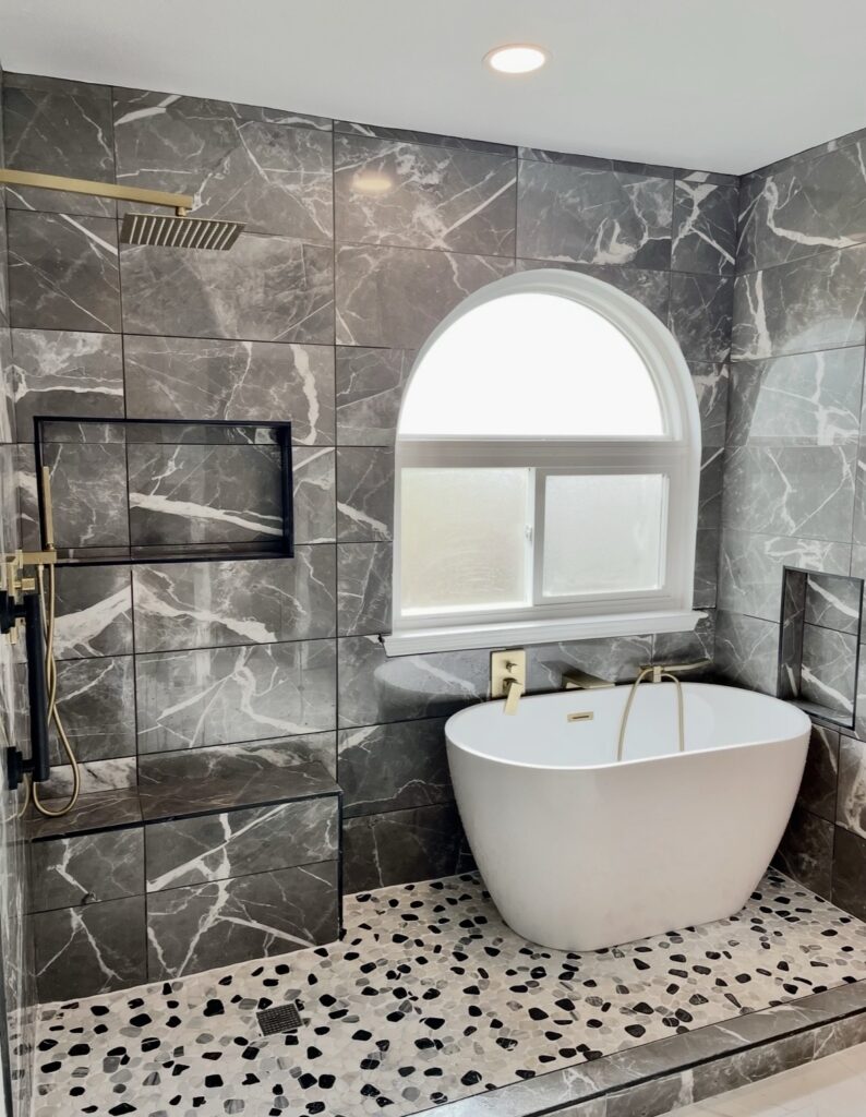 Freestanding white bathtub beside a shower with dark marble-look tile, a recessed niche, and pebble-pattern flooring.