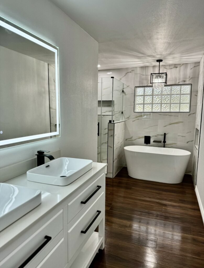 White bathroom vanity with vessel sinks, illuminated mirror, freestanding tub, glass shower and black fixtures.