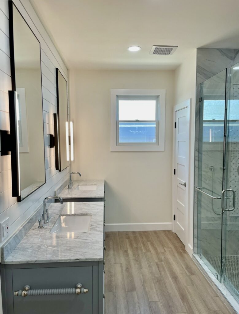 Double vanity with gray cabinetry, light countertops, black-framed mirrors and a glass shower.