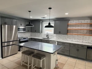 Gray kitchen cabinets, a white subway tile backsplash, floating wood shelves and a contrasting island with black pendant lights.
