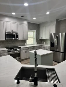 Light gray kitchen cabinets, patterned tile backsplash, a small island and a black sink with a spring faucet.
