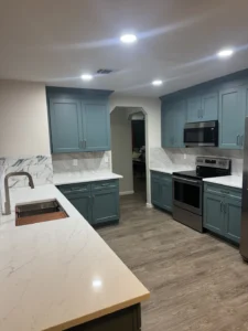 Teal kitchen cabinets, light veined counters and backsplash, stainless steel appliances and a wood-look floor.