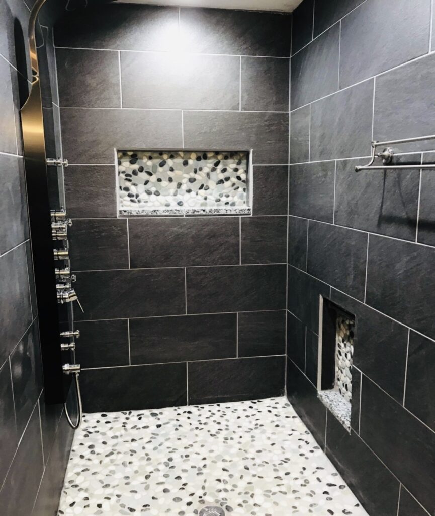 Finished shower with dark rectangular wall tile, a mosaic niche, glass enclosure and pebble-pattern floor.